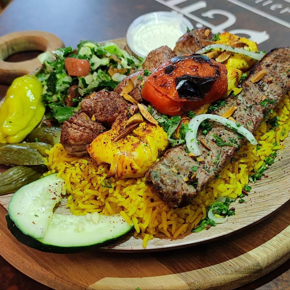 A vibrant Mediterranean platter with grilled meats, yellow rice, salad, pickles, and a side of sauce.