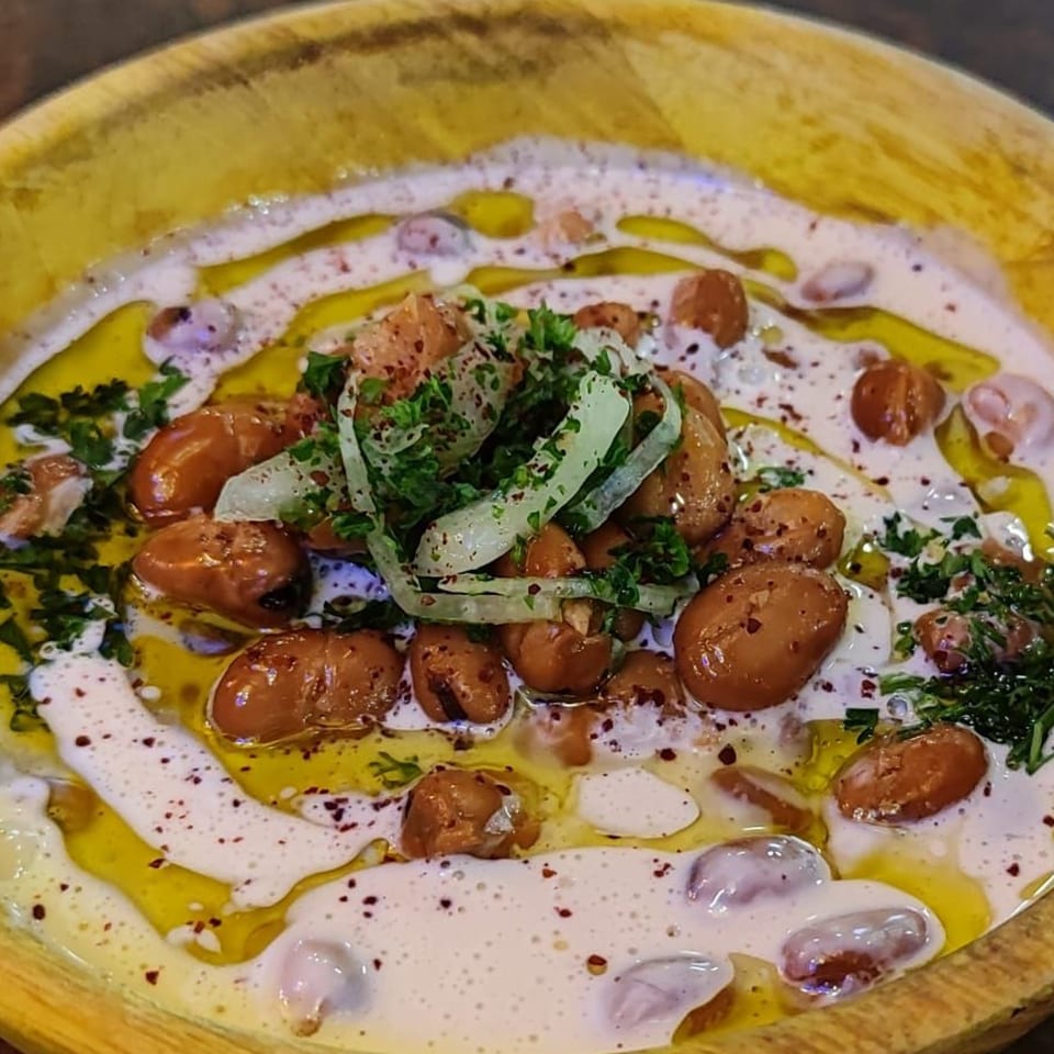 Foul Mudammas, a Mediterranean dish with fava beans, tahini, olive oil, parsley, and onions in a wooden bowl.